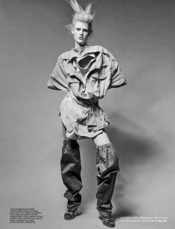 Fashion shot of a female model in a light-colored shirt dress and thigh-high boots standing against a grey studio backdrop with bright lighting.