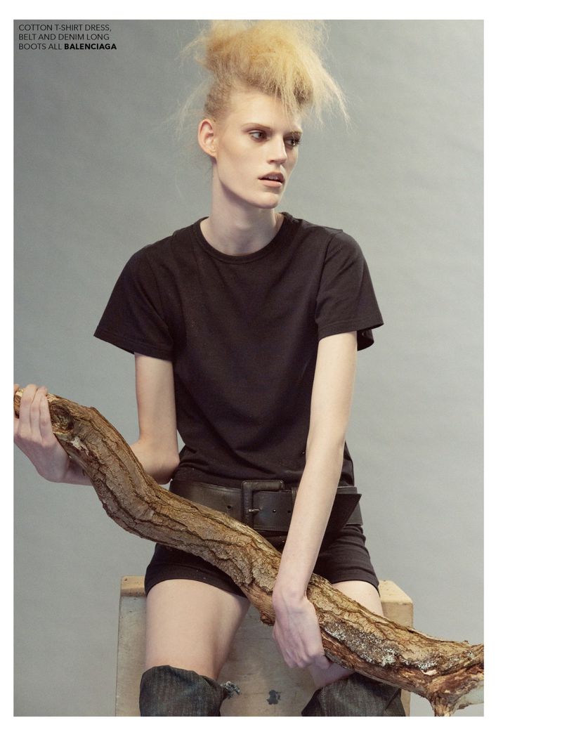 Fashion editorial shot of a blonde model in dark casual clothing, holding a large branch in a grey indoor studio environment.