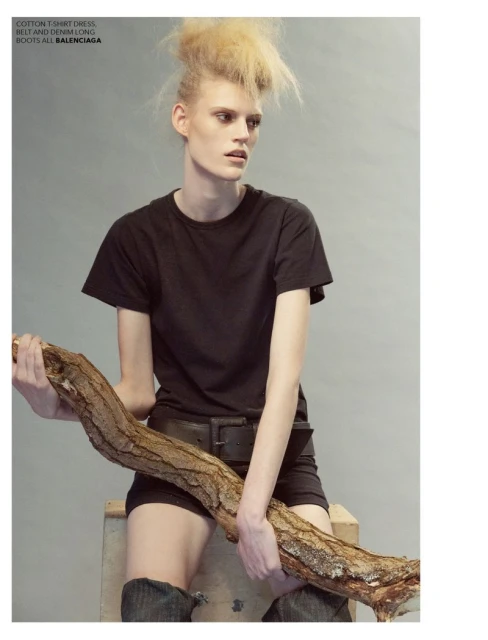 Fashion editorial shot of a blonde model in dark casual clothing, holding a large branch in a grey indoor studio environment.