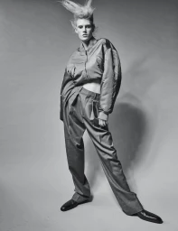 Model with spiky hair wearing a cropped jacket and trousers against a grey seamless studio background.