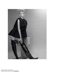 Black and white portrait of a female model wearing a layered black dress and tall leather boots against a grey studio backdrop.