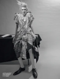 Model Bodine Van Galen seated wearing a patterned lace dress and black leather shoes against a grey background.