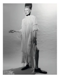 Monochrome fashion editorial image of model Bodine Van Galen wearing a pleated white dress and tall black hat against a grey studio backdrop.