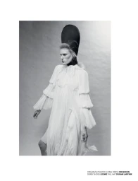 Model Bodine Van Galen in a white organza pleated long dress and tall black hat against a grey seamless studio background.