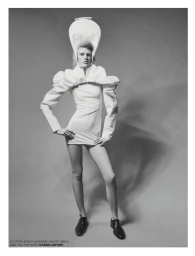 Model Bodine Van Galen in a white structured mini dress and tall sculptural hat against a grey studio backdrop.