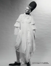 Model in white pleated dress and tall black hat posing against a seamless grey backdrop for Re-Edition Magazine.