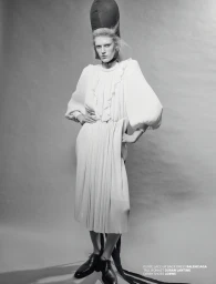 Monochrome fashion portrait of a model wearing a ruffled white dress and tall structured bonnet against a grey background.