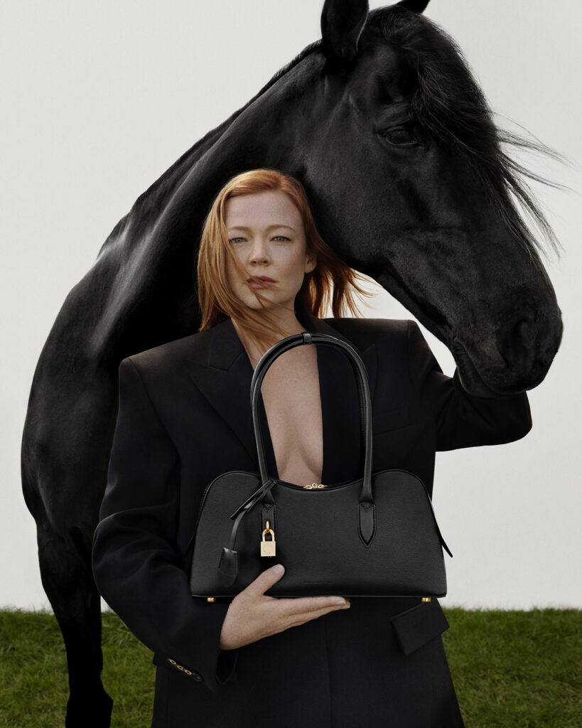 Sarah Snook poses in a black blazer with a black leather handbag and a black horse, set against a blurred green and white backdrop.