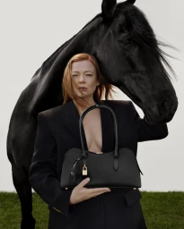 Sarah Snook wearing a black blazer holding a black handbag with a black horse behind her against a plain background