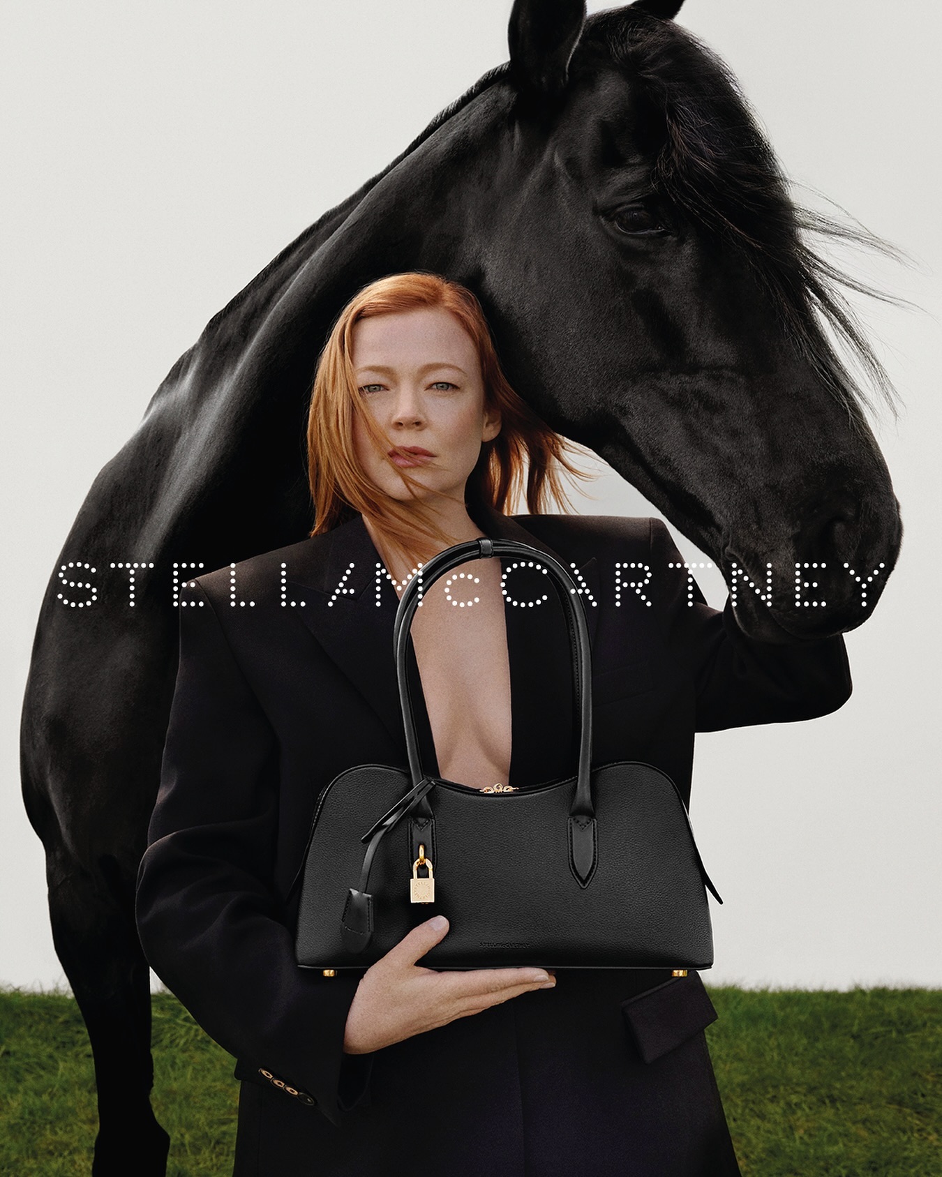 Sarah Snook poses with a black horse and a black handbag by Stella McCartney, wearing a black suit, against a bright background.