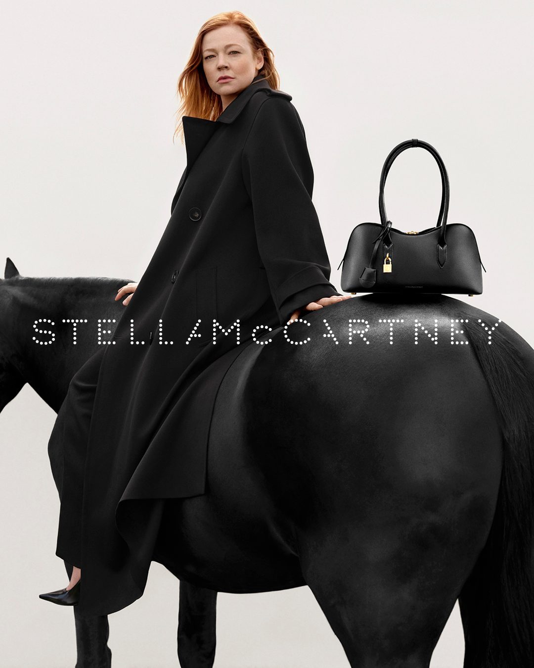 Sarah Snook sits astride a black horse wearing a black coat, with a Stella McCartney black bag resting on the horse's back. 