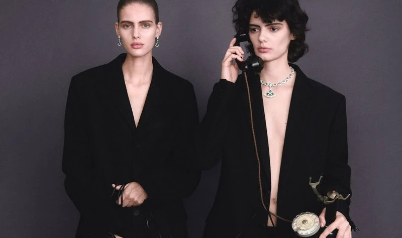 Two women pose in black jackets against a gray backdrop. One woman is on the phone, accessorized with statement necklace and earrings.