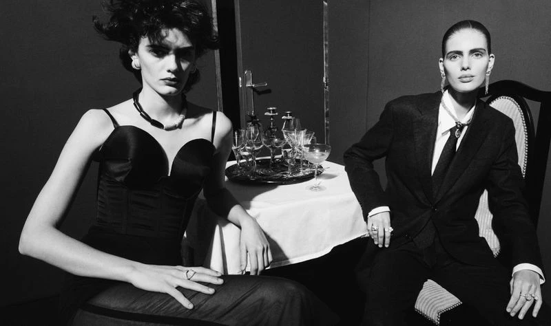 Black and white studio shot of two adult women at a table wearing fashionable formalwear. One woman is wearing a suit and tie.