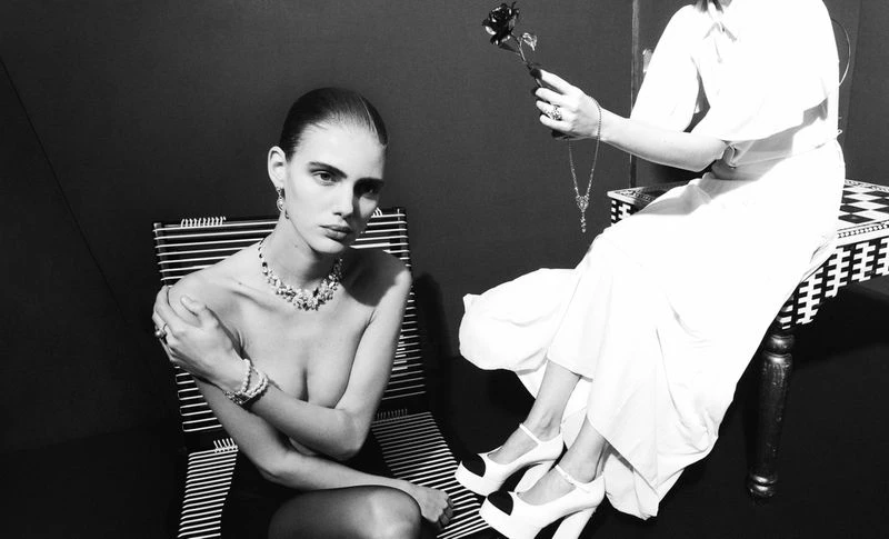 Black and white portrait of a young adult woman with jewelry seated next to another woman offering a black rose in a studio setting.