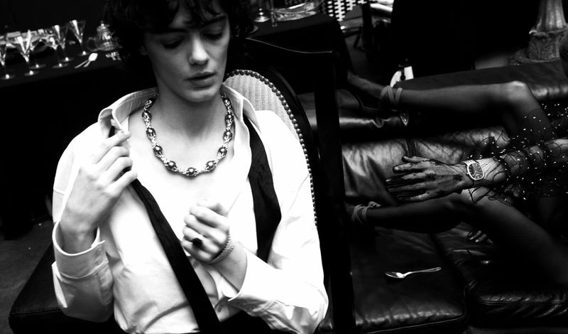 Monochrome portrait of a woman in a white shirt and necklace, seated in chair with a woman reclining on a sofa in the background.