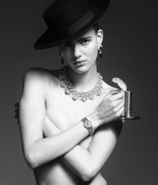 Monochrome fashion portrait of a topless adult female with jewelry and a black hat holding a quill pen with a neutral expression.