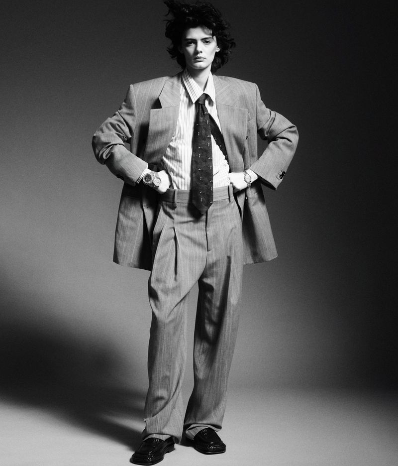 A full-length black-and-white fashion portrait of a female model wearing an oversized pinstripe suit, tie, and loafers.