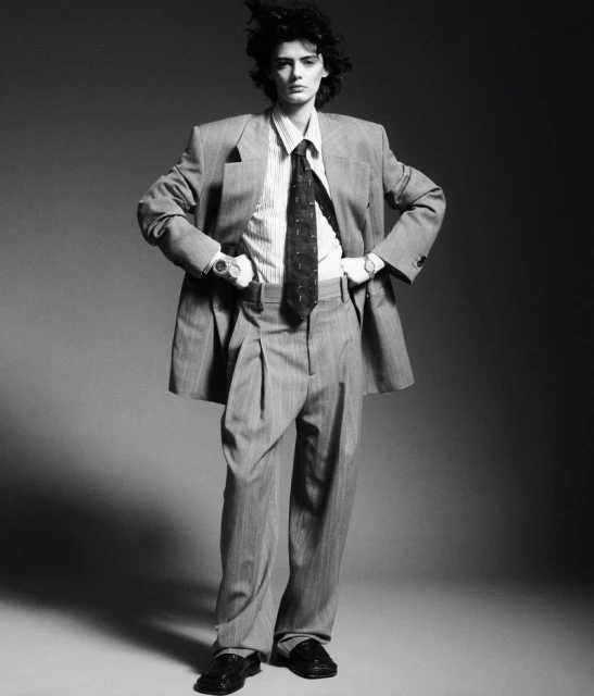 A full-length black-and-white fashion portrait of a female model wearing an oversized pinstripe suit, tie, and loafers.