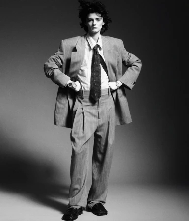 A full-length black-and-white fashion portrait of a female model wearing an oversized pinstripe suit, tie, and loafers.