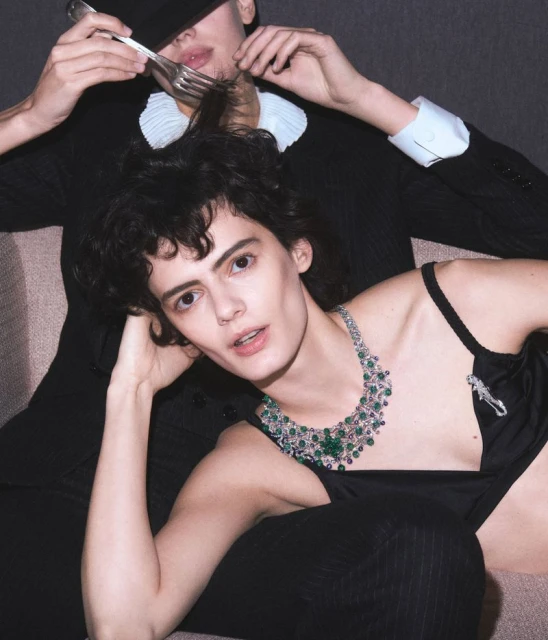 Two fashion models pose in a studio. One wears a black suit and the other wears a necklace and lingerie top with pinstripe pants.