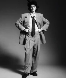 Model in oversized grey suit with striped shirt and dark tie posing against a neutral dark gradient background