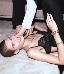Model lying on a white textured rug wearing a black lace bralette with another person standing over her