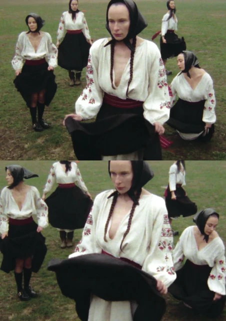 A group of women in traditional dress stand outdoors, wearing white embroidered shirts, black skirts, and headscarves on a grassy field.