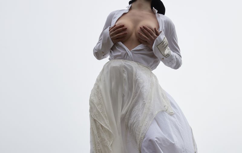 A woman in a white shirt and skirt covers her breasts with her hands in an outdoor setting with a bright background, conveying a sense of modesty.