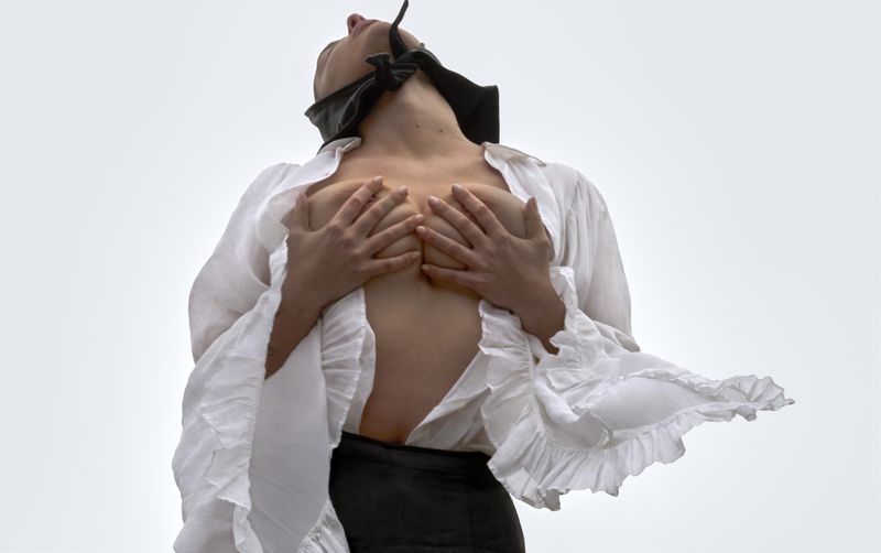 Artistic portrait of a Caucasian woman with a blindfold, wearing a white flowing ruffled shirt, eyes up, against a pale grey background.