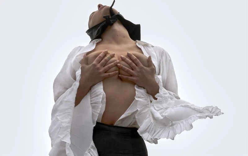Artistic portrait of a Caucasian woman with a blindfold, wearing a white flowing ruffled shirt, eyes up, against a pale grey background.