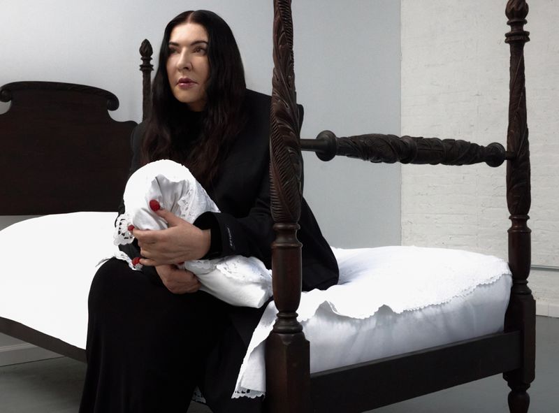 Woman in black sits on antique four-poster bed holding white lace pillow in white room. Dark carved wood contrasts white linens.