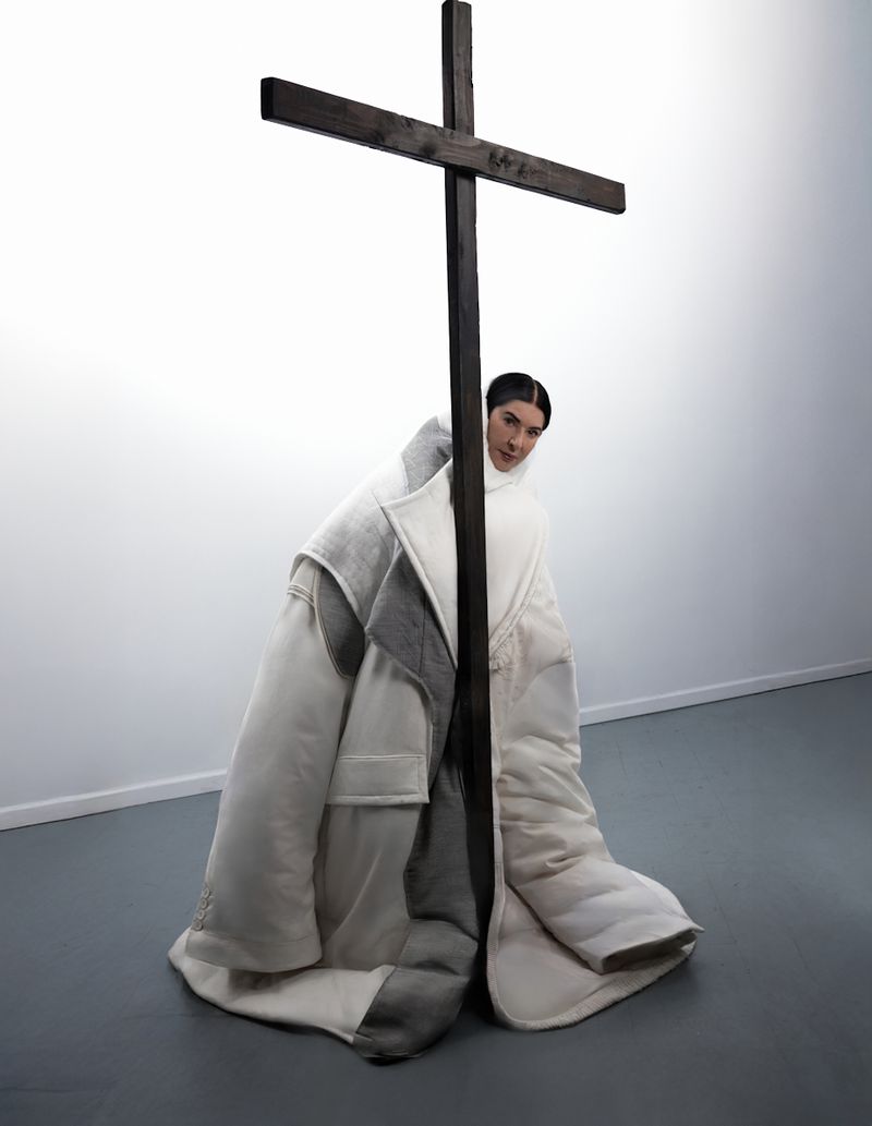 Woman posing in oversized white coat with a large wooden cross in an indoor photo studio with white walls and a grey floor.
