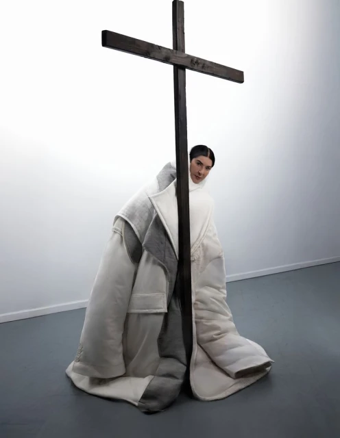 Woman posing in oversized white coat with a large wooden cross in an indoor photo studio with white walls and a grey floor.