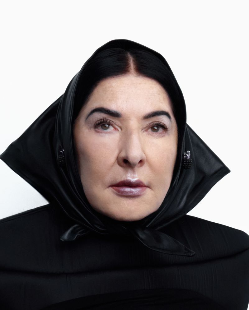 Portrait of a mature woman with dark hair and glossy lips wearing a black headscarf and a black dress against a white background.