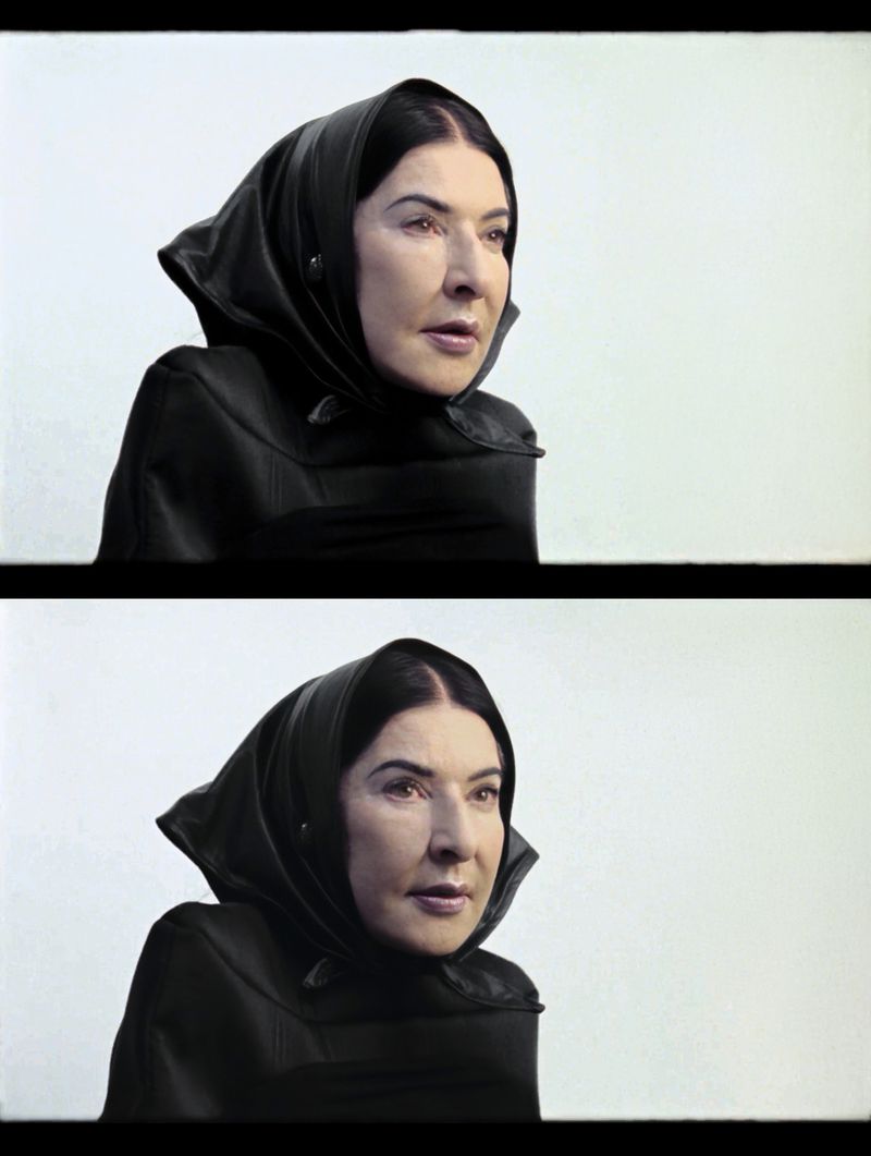 Portrait of an adult woman with dark hair wearing a black headscarf and jacket against a plain white background. Studio lighting.