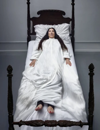 Overhead shot of a woman lying on a dark wood, four-poster bed, covered by a white, vintage-style bedspread and pillow in a simple bedroom.
