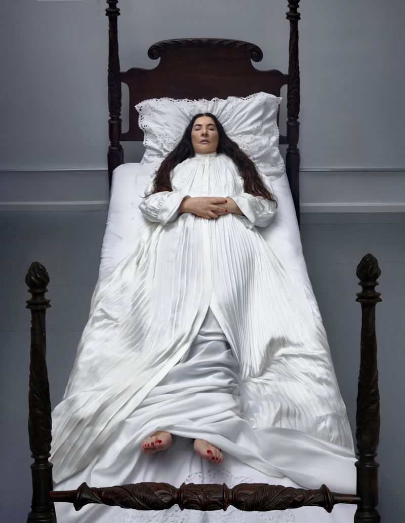Adult woman in a white dress reclines on a wooden four-poster bed in a brightly lit room with her hands folded on her chest.