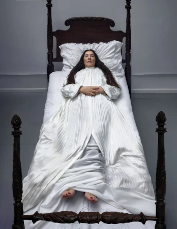 Adult woman in a white dress reclines on a wooden four-poster bed in a brightly lit room with her hands folded on her chest.