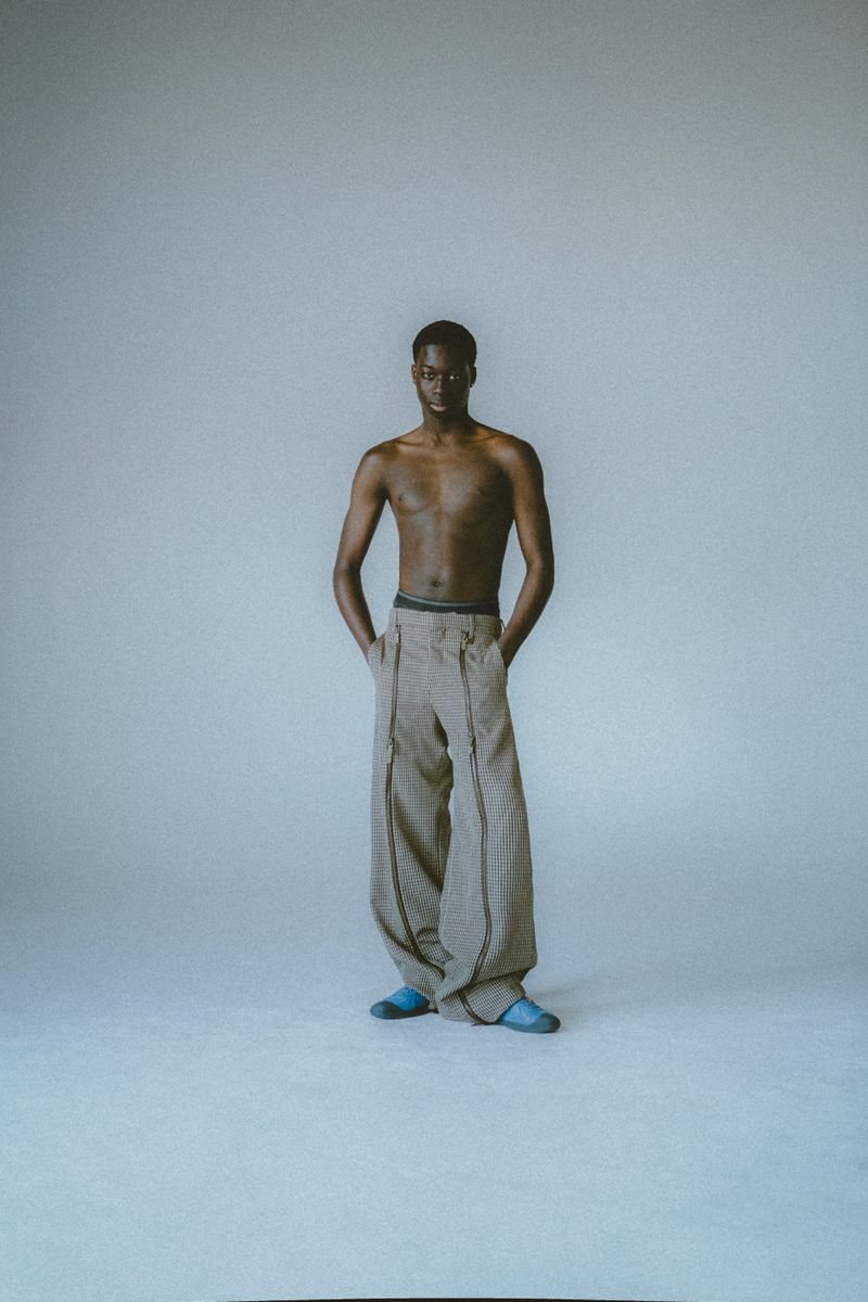 Man in oversized pants and slippers stands in front of white background with hands in pockets, photographed in full-length in a studio.