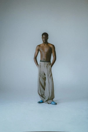 Man in oversized pants and slippers stands in front of white background with hands in pockets, photographed in full-length in a studio.