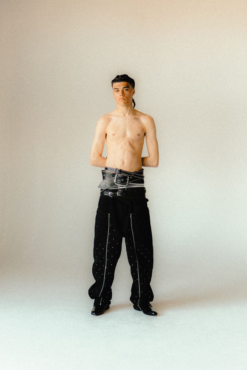 A shirtless man stands in front of a plain white background wearing black pants and multiple belts. The subject is well-lit.