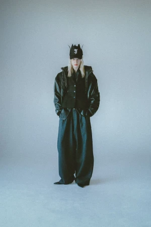 Full-length shot of a person with long blonde hair wearing a baseball cap, vest, jacket, and loose pants against a grey background.