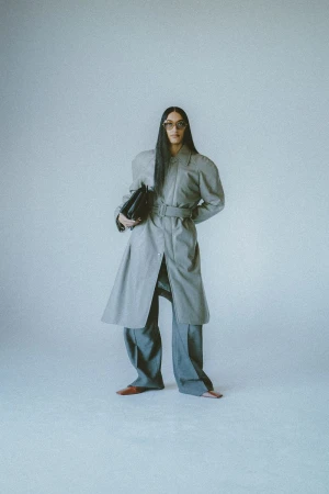 A woman in a grey belted trench coat and wide-leg pants holds a black handbag against a grey studio backdrop, showcasing modern fashion.