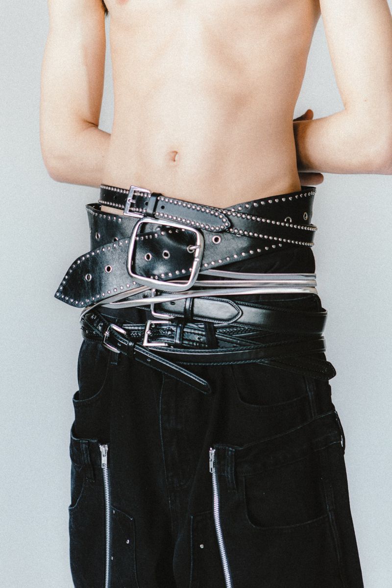 Torso shot of a person wearing black pants with zipper details and multiple black belts wrapped around their waist in a studio setting.