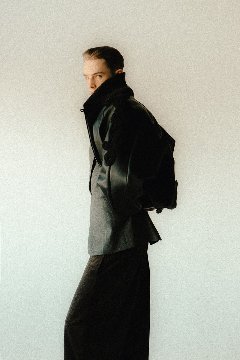 An adult with short hair wearing a black jacket over a gray shirt and a long black skirt stands against a pale background.
