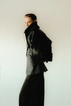 An adult with short hair wearing a black jacket over a gray shirt and a long black skirt stands against a pale background.