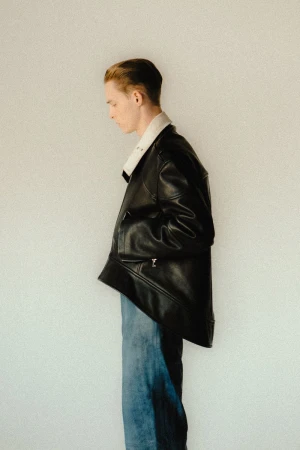 Man in a black leather shearling jacket and blue jeans poses in front of a white background in a studio, side profile.