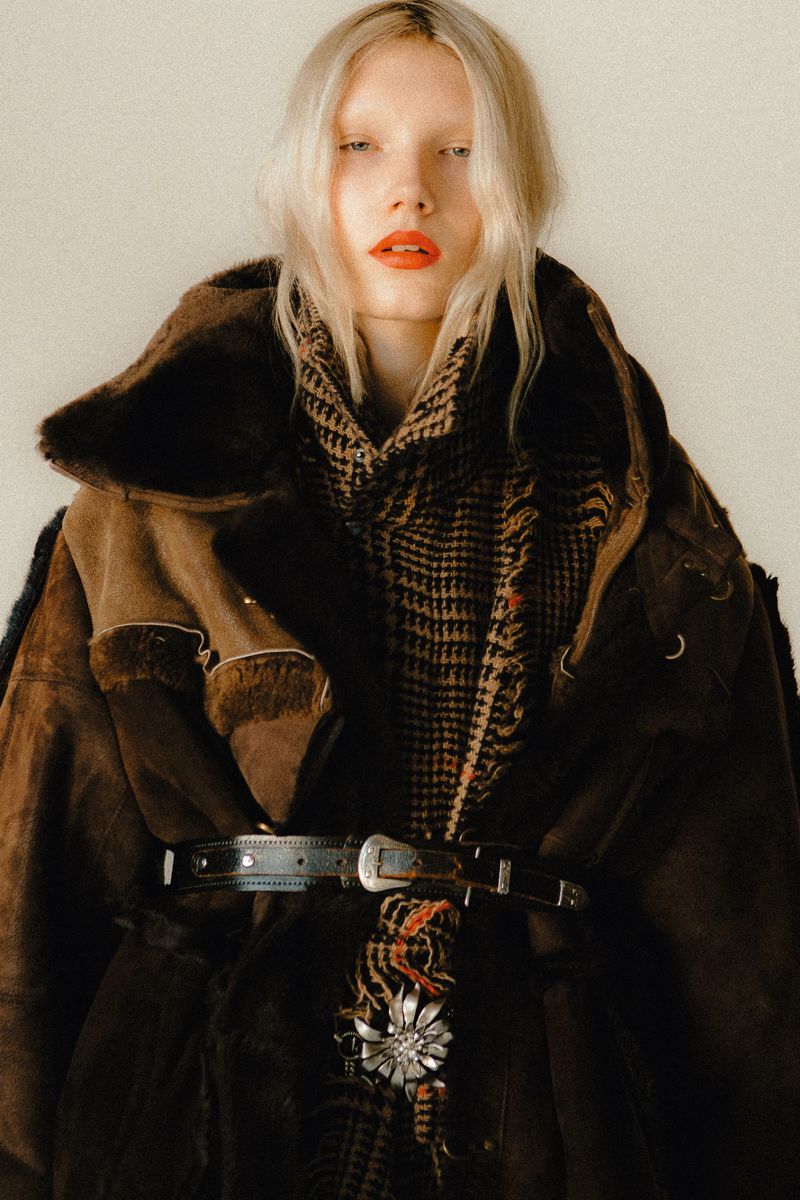 Portrait of a blonde woman wearing a brown suede coat with fur collar, scarf, and silver belt, posing against an off-white background.
