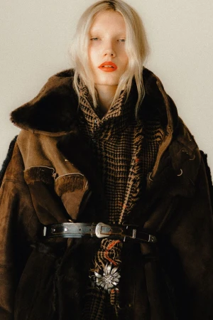 Portrait of a blonde woman wearing a brown suede coat with fur collar, scarf, and silver belt, posing against an off-white background.
