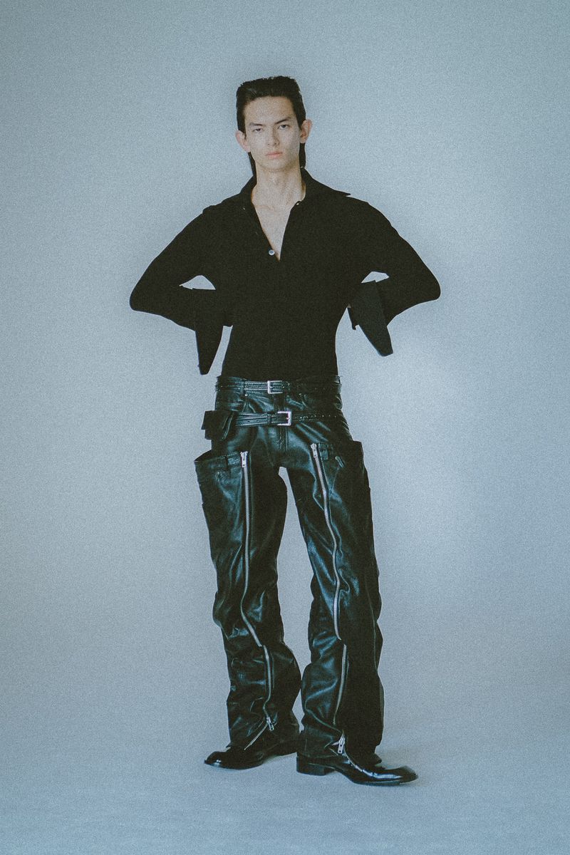 A slender male model poses in a long-sleeved black shirt and shiny black pants with silver zippers against a solid grey background.